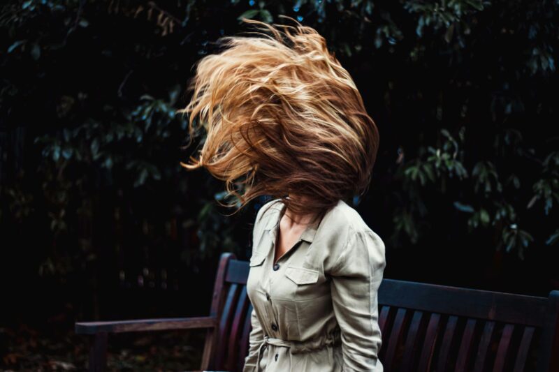 A dynamic photo of a woman flipping her hair while sitting on a park bench outdoors.