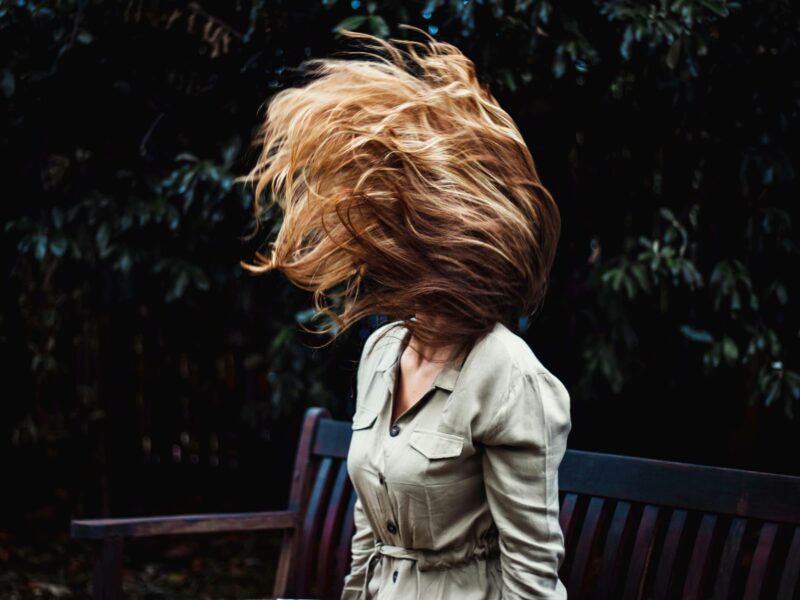 A dynamic photo of a woman flipping her hair while sitting on a park bench outdoors.